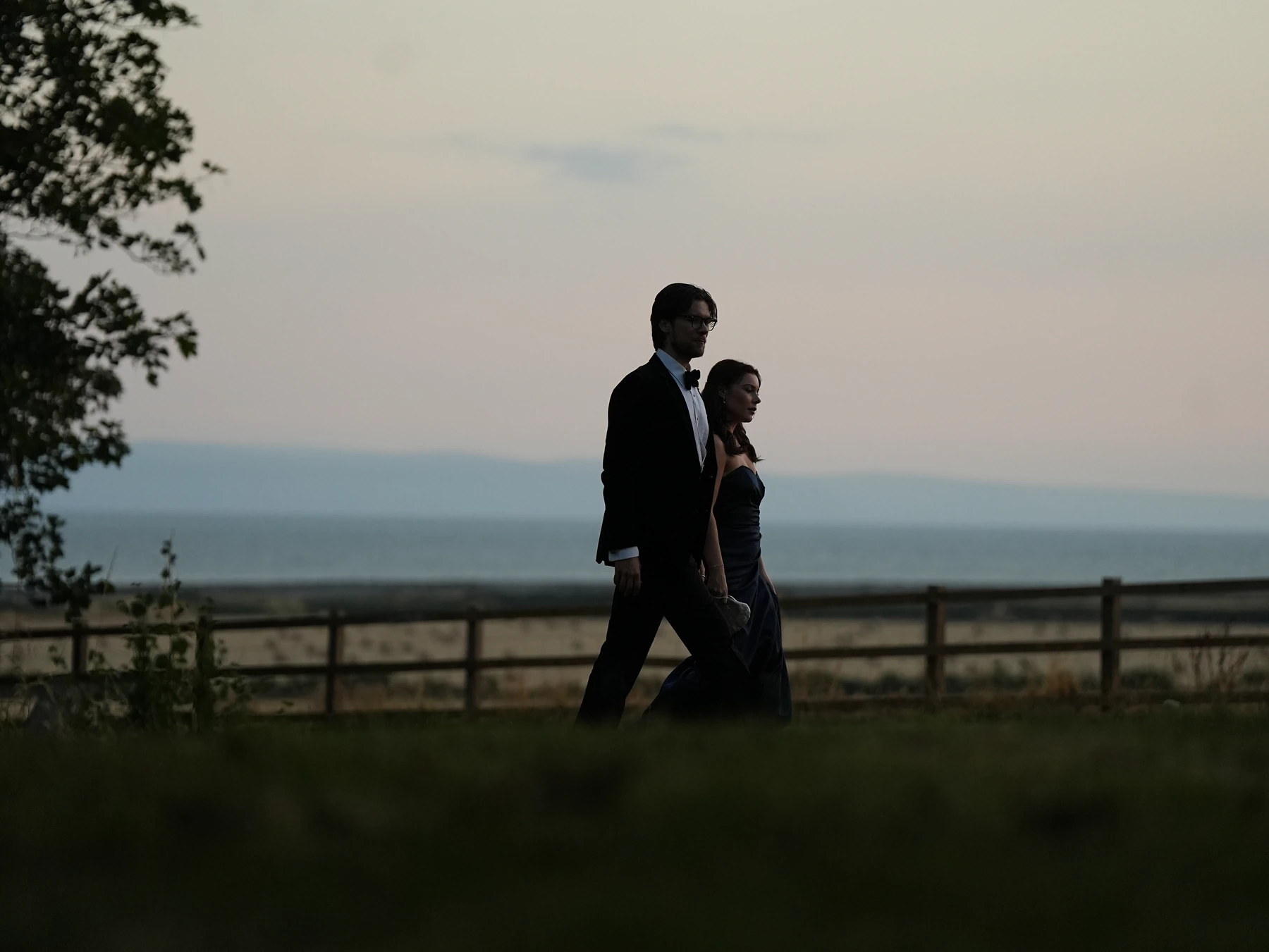 Two cast members in evening wear walking beside the coast at dusk
