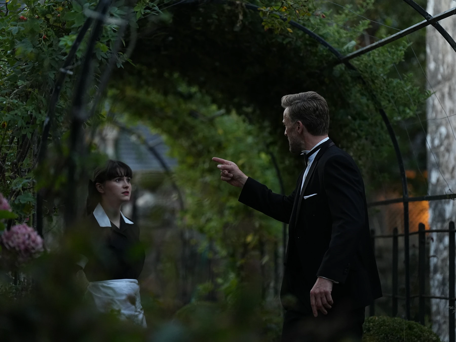 A man in a dinner suit speaking to a maid beneath a leafy garden archway