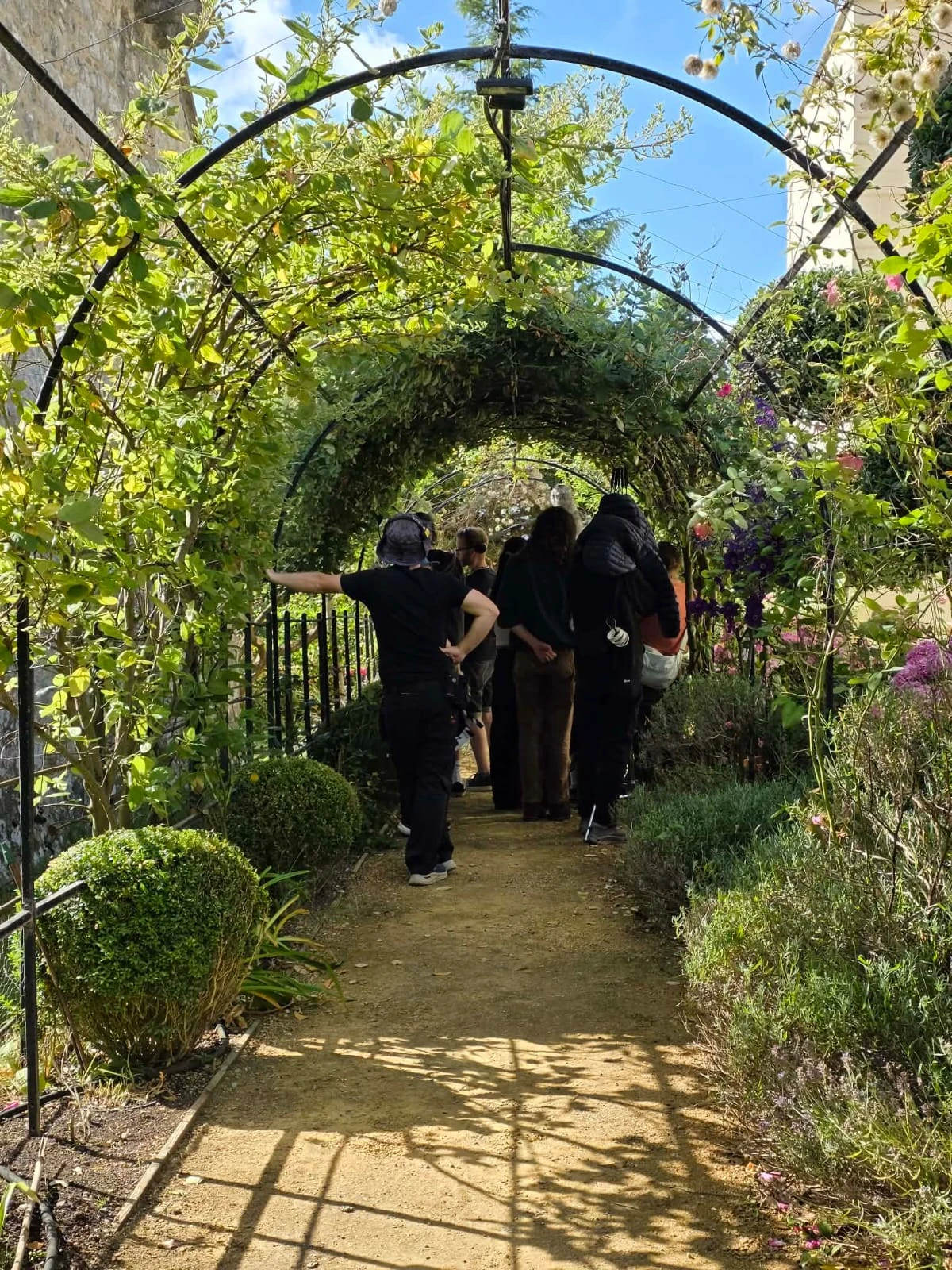 Crew preparing a scene beneath a leafy garden archway in the sunshine