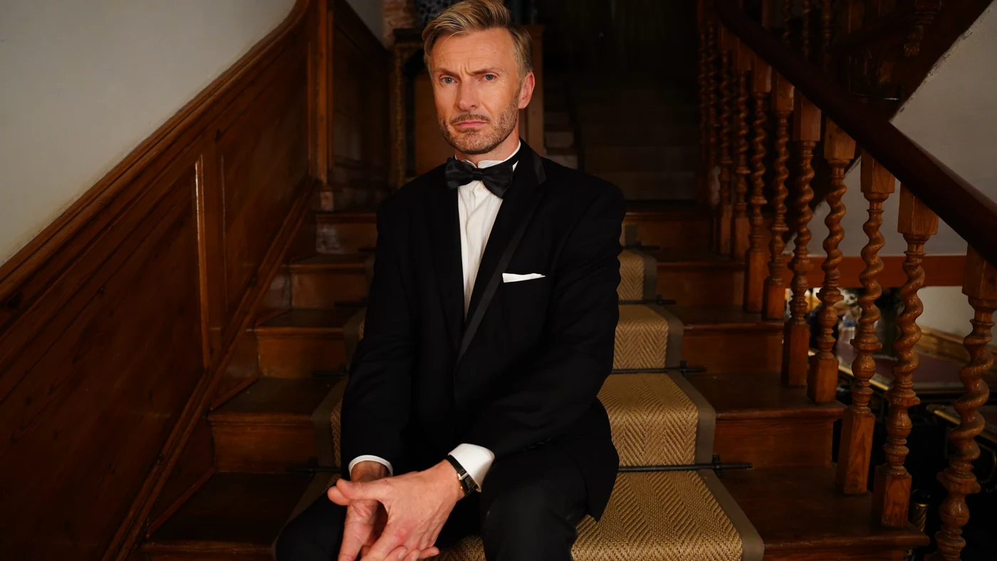 Simon Price in a black tuxedo seated on a staircase